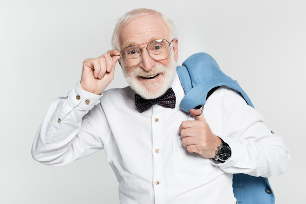 Positive senior man in bow tie holding eyeglasses and jacket isolated on grey  - 写真・画像