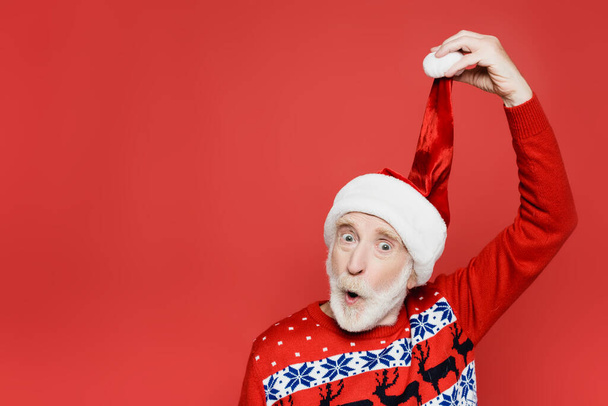 Excited elderly man holding santa hat isolated on red - Фото, зображення