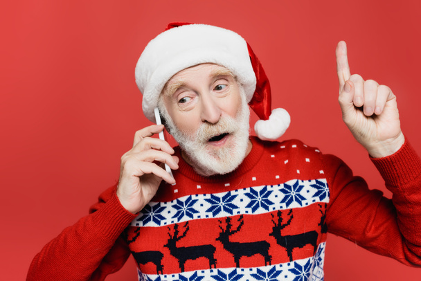 Senior man in santa hat talking on smartphone and pointing with finger isolated on red - Foto, Bild