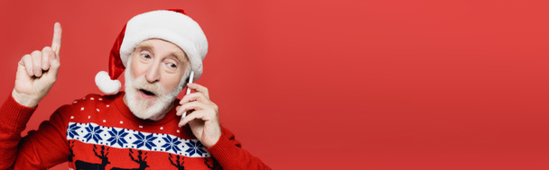 Starší muž v Santa klobouku mluví na smartphonu a ukazuje prstem izolovaným na červenou, banner - Fotografie, Obrázek