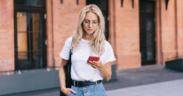 Attractive caucasian blonde female in eyewear reading income message on smartphone standing on urban setting background, hipster girl searching location via mobile phone application on street - Foto, Bild