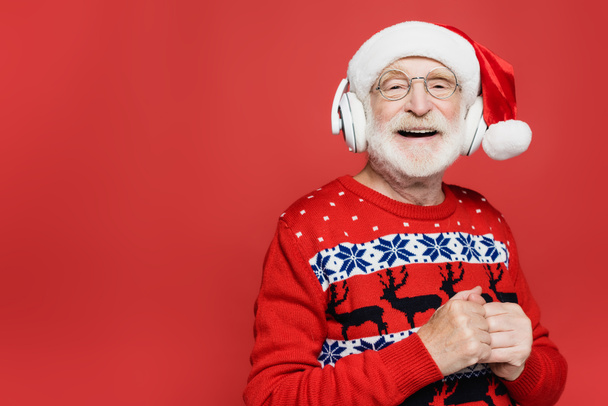 Positive senior man in santa hat and sweater listening music in headphones isolated on red - 写真・画像