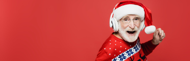 Cheerful senior man in santa hat and headphones dancing isolated on red, banner  - Foto, imagen
