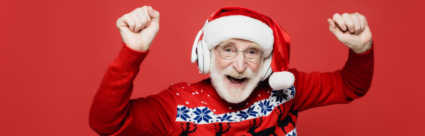 Veselý starší muž v Santa klobouku tanec při poslechu hudby ve sluchátkách izolovaných na červené, banner  - Fotografie, Obrázek