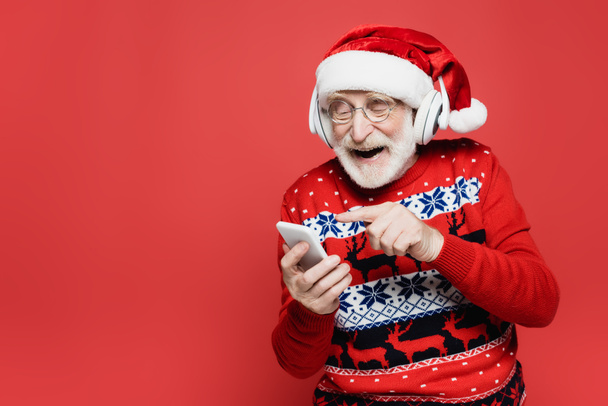 Kulaklıklı ve Noel Baba şapkalı gülümseyen yaşlı adam kırmızıda izole edilmiş akıllı telefonu işaret ediyor. - Fotoğraf, Görsel