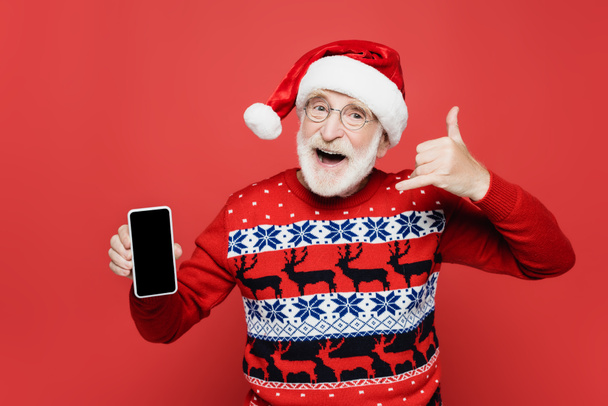 Fröhlicher Senior mit Weihnachtsmütze und Pullover zeigt Smartphone mit leerem Bildschirm und gestikuliert isoliert auf Rot - Foto, Bild