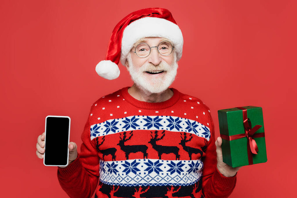 Lächelnder älterer Mann mit Weihnachtsmannmütze, Smartphone mit leerem Bildschirm und Geschenk auf rotem Grund - Foto, Bild