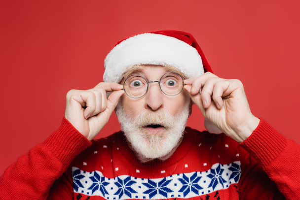 Shocked senior man in santa hat and eyeglasses looking at camera isolated on red  - Фото, изображение