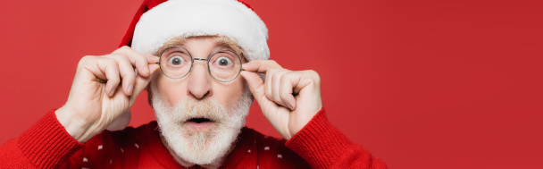 Überraschter Senior mit Weihnachtsmütze und Brille, der auf rotem Banner isoliert in die Kamera blickt  - Foto, Bild