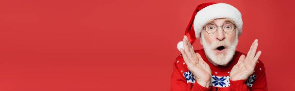 Shocked elderly man in santa hat and sweater looking at camera isolated on red, banner - Photo, image