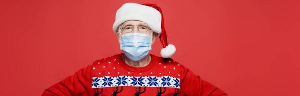 Senior man in santa hat and medical mask looking at camera isolated on red, banner  - Foto, Imagem