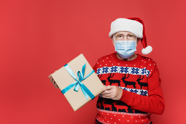 Senior man in medical mask and santa hat holding present with bow isolated on red  - Foto, Bild