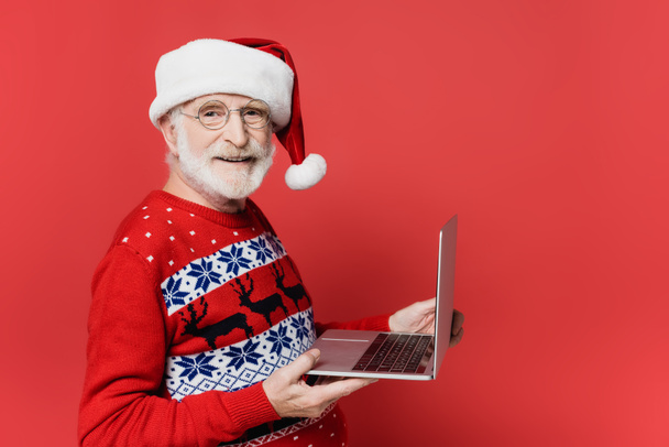 glimlachende oudere man in santa hoed met laptop op rode achtergrond - Foto, afbeelding
