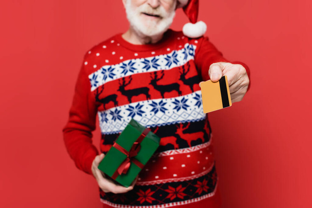 Przycięty widok karty kredytowej w ręku starszego mężczyzny w czapce Mikołaja z teraźniejszością na niewyraźnym pierwszym planie na czerwonym tle - Zdjęcie, obraz