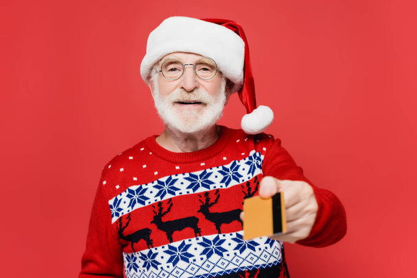 Smiling elderly man in santa hat holding credit card on blurred foreground isolated on red  - Foto, Imagen