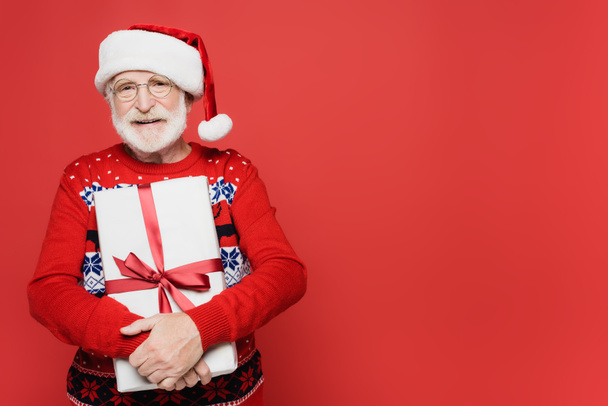 Elderly man in santa hat smiling while holding gift isolated on red  - Photo, Image