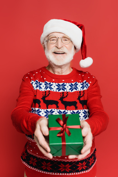 Gülümseyen son sınıf öğrencisi, Noel Baba şapkası ve örgü süveter giyerek kırmızı üzerine izole edilmiş hediye kutusu gösteriyor.  - Fotoğraf, Görsel