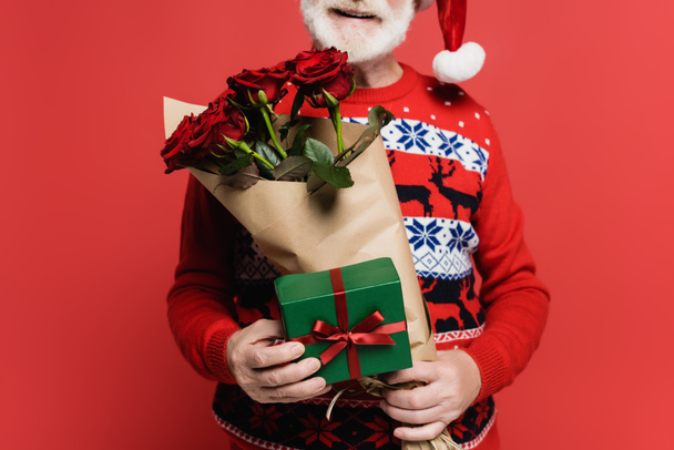 Vista cortada de homem sênior sorridente em santa chapéu segurando caixa de presente e buquê de rosas isoladas em vermelho  - Foto, Imagem