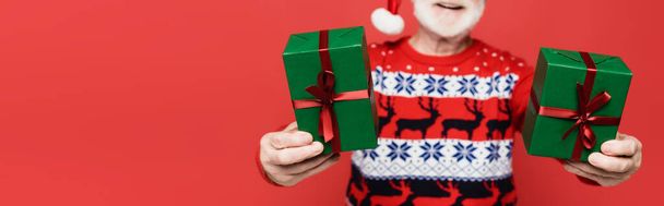 Cropped view of gifts in hands of senior man in santa hat on blurred background isolated on red, banner  - Fotó, kép