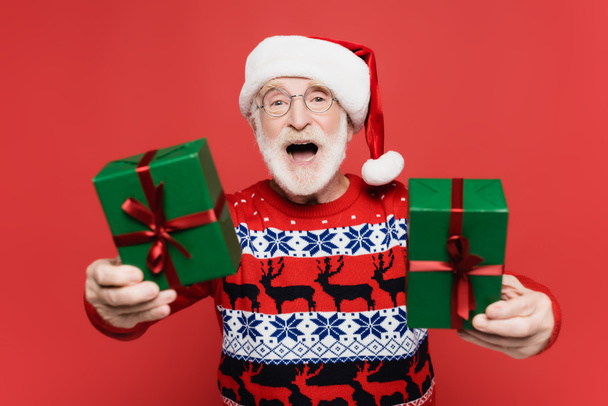 Cheerful elderly man in santa hat holding gift boxes on blurred foreground isolated on red  - Fotó, kép