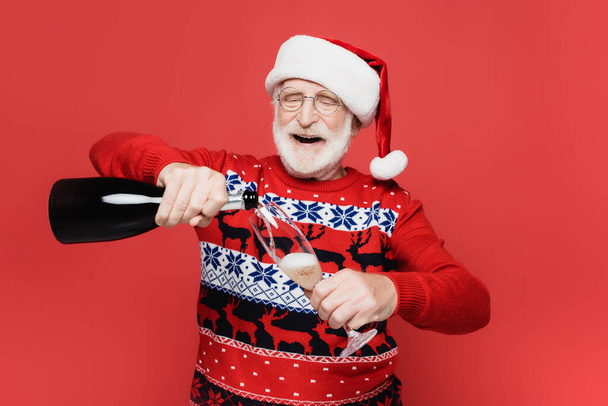 Smiling senior man in santa hat pouring champagne in glass isolated on red  - Zdjęcie, obraz