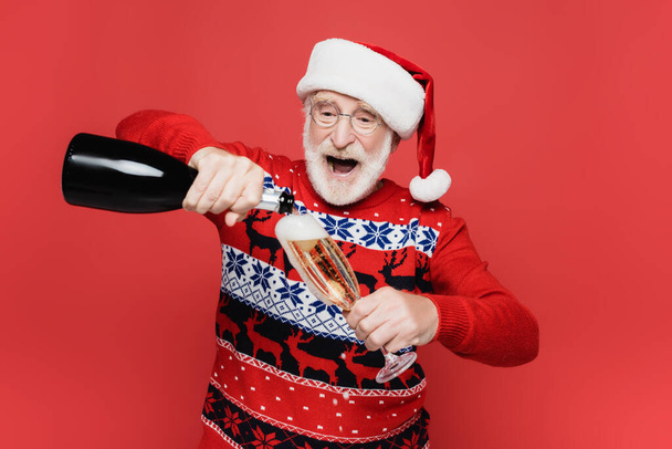 Excited elderly man in santa hat pouring champagne in glass isolated on red  - Foto, immagini