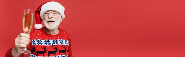 Uomo anziano sorridente in cappello di Babbo Natale che tiene un bicchiere di champagne in primo piano sfocato isolato su rosso, striscione  - Foto, immagini