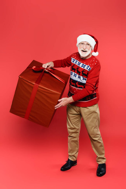 Smiling elderly man in santa hat holding present with bow and looking at camera on red background - Foto, Bild