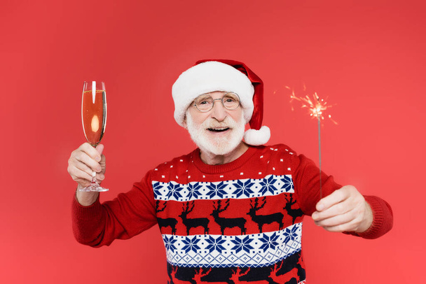 Smiling senior man in santa hat holding glass of champagne and sparkler isolated on red  - Фото, изображение