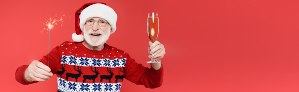 Elinde şampanya kadehi ve kırmızı pankartta izole edilmiş maytap tutan Noel Baba şapkalı gülümseyen yaşlı adam.  - Fotoğraf, Görsel