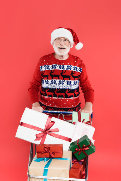 Positieve oudere man in kerstmuts kijkt weg in de buurt van cadeautjes in winkelwagen geïsoleerd op rood  - Foto, afbeelding