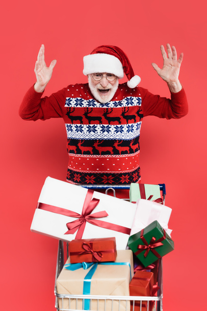 Захоплений старший чоловік у капелюсі Санта дивиться на подарунки в кошику для покупок ізольовано на червоному
  - Фото, зображення