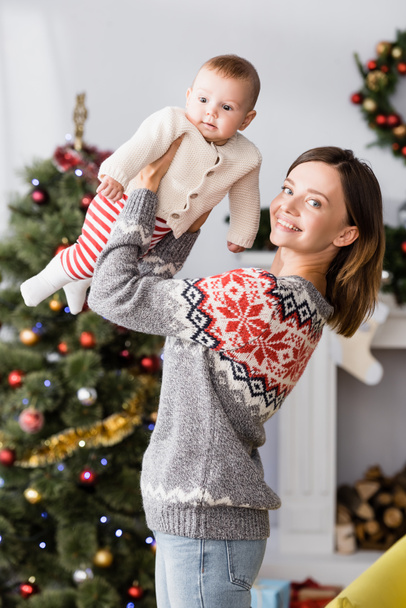 happy woman holding in arms baby boy near blurred christmas tree on background - Фото, зображення