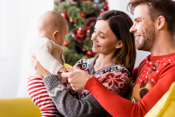 背景にぼやけたクリスマスツリーの近くにいる赤ちゃんの息子を見ている陽気な両親 - 写真・画像