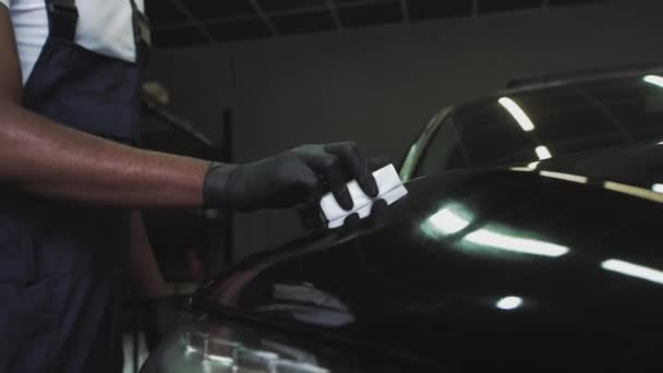 Ένας Αφρο-Αμερικανός υπάλληλος εφαρμόζει ένα προστατευτικό στρώμα. Εφαρμογή προστασίας για προβολείς. Επαγγελματική Car Κεραμικά Εργαζόμενος εφαρμόζει ένα στρώμα κεραμικών προστατευτικό κάλυμμα βροχής στα παράθυρα του αυτοκινήτου - Πλάνα, βίντεο