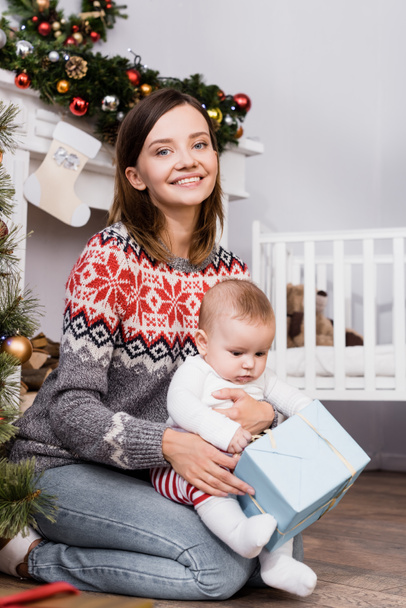 mujer alegre sosteniendo presente cerca de bebé niño en apartamento decorado - Foto, imagen