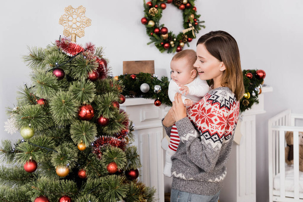 sonriente mujer sosteniendo en brazos bebé niño cerca decorado árbol de Navidad - Foto, Imagen