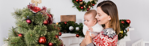 donna sorridente che tiene in braccio il bambino vicino all'albero di Natale decorato, striscione - Foto, immagini