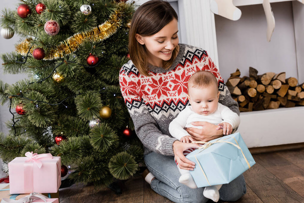 smiling woman holding present near infant son and decorated christmas tree - Foto, afbeelding