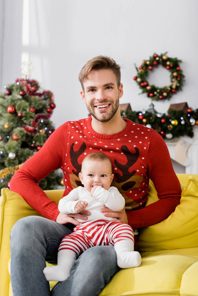cheerful father sitting with baby boy on sofa with christmas tree on blurred background - Zdjęcie, obraz