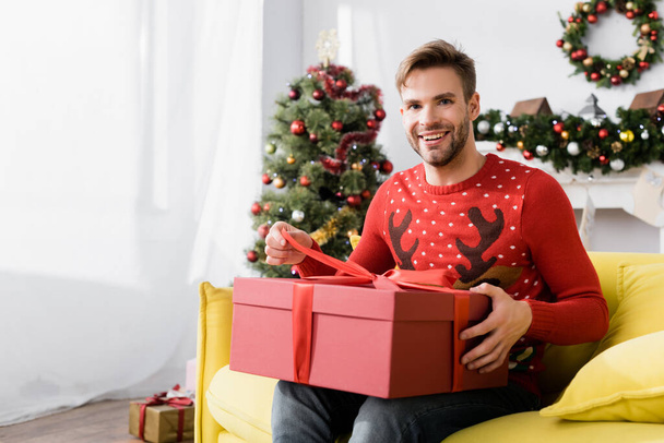 glücklicher Mann in rotem Pullover mit eingewickeltem Geschenk, während er auf dem Sofa sitzt, mit verschwommenem Weihnachtsbaum im Hintergrund  - Foto, Bild