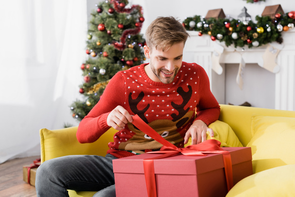 homme souriant en pull rouge tenant ruban sur cadeau enveloppé tout en étant assis sur le canapé avec arbre de Noël flou sur le fond  - Photo, image