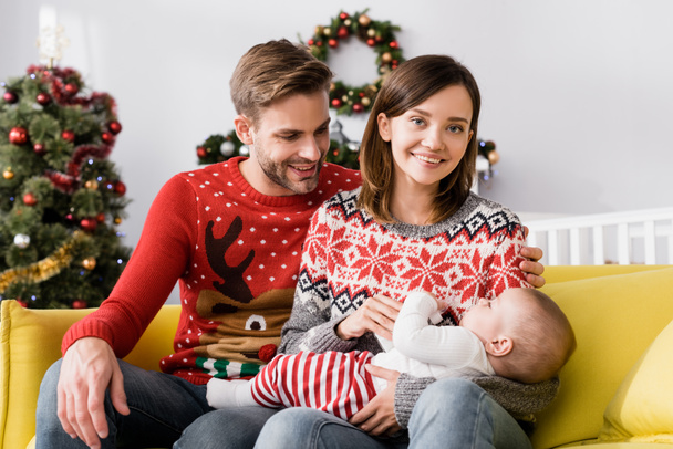 heureux parents en chandails souriant près du fils de bébé avec arbre de Noël sur fond flou  - Photo, image