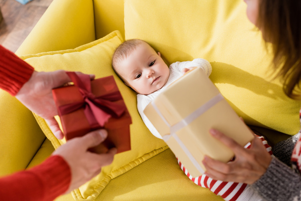 Säugling auf Sofa liegend und Eltern mit verpackten Weihnachtsgeschenken im verschwommenen Vordergrund anschauend  - Foto, Bild