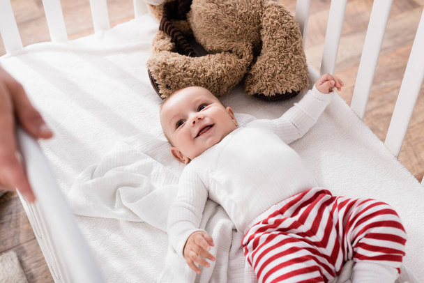 happy infant boy lying in baby crib and looking at father on blurred foreground  - Foto, afbeelding