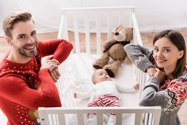 high angle view of happy parents looking at camera near baby boy in crib on blurred background  - Fotografie, Obrázek