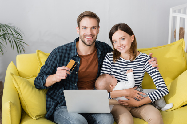 счастливый мужчина держит кредитную карту рядом с женой с младенцем сыном и ноутбуком  - Фото, изображение