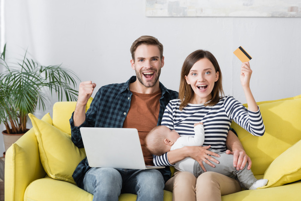 cheerful woman with infant son holding credit card near excited husband while online shopping  - Fotoğraf, Görsel