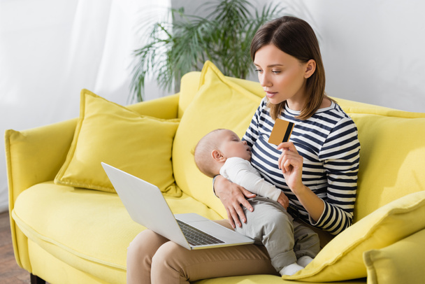 Frau mit Säugling hält Kreditkarte neben Laptop, während sie auf Sofa sitzt - Foto, Bild