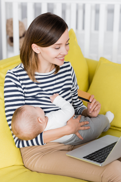 smiling woman with infant son holding credit card near laptop while sitting on sofa - Foto, imagen